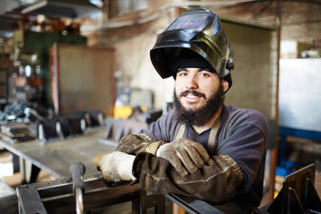 Smiling handsome young craftsman in welding helmet leaning on constriction frame and looking at cameraの写真素材