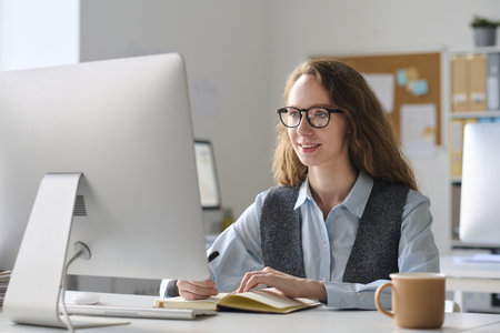 Young businesswoman in eyeglasses working on computer at her workplace and making notes in notepadの写真素材