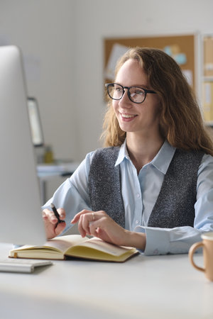 Vertical image of smiling young woman using computer at her work while sitting at her workplace with notepadの写真素材