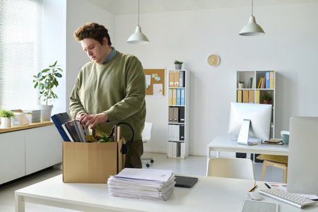 Young office worker standing at his workplace and packing his things in box after layoffの写真素材
