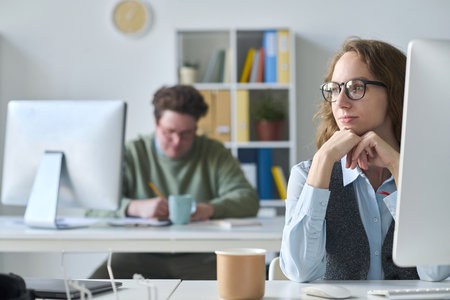 Young businesswoman in eyeglasses sitting at her workplace with computer with her colleague working in backgroundの写真素材