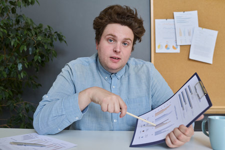 Portrait of young businessman looking at camera while sitting at table and examining financial documents in officeの写真素材