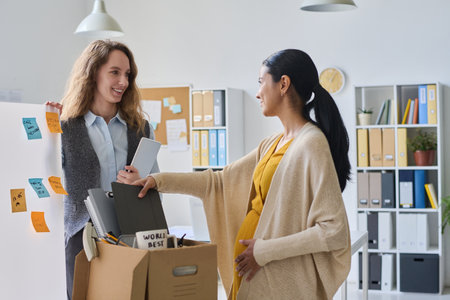 Businesswoman saying goodbye to her pregnant colleague while she packing things in box, they standing in office and talkingの写真素材