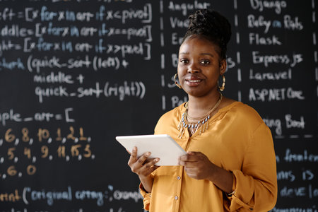 Portrait of African American teacher smiling at camera while using digital tablet at lesson standing against blackboardの写真素材