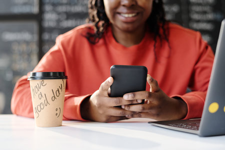 Close-up of African American teacher using smartphone while sitting at table with coffee drink and laptop in the classroomの写真素材