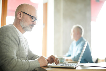 Serious concentrated senior male freelancer with beard and mustache sitting at table and working with laptop in public placeの写真素材