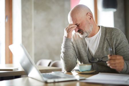 Tired bald senior male freelancer with beard rubbing eyes while taking off glasses in cafeの写真素材