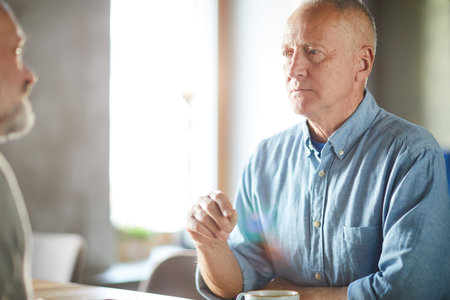 Serious pensive handsome mature man in blue shirt sitting at table and communicating with friend in modern cafeの写真素材