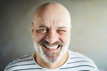 Portrait of excited emotional bald mature man with gray beard and mustache laughing cheerfully and winking at cameraの写真素材