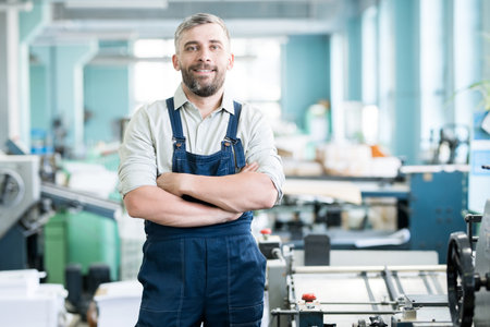 Cheerful content confident bearded typography worker in overall crossing arms on chest and looking at camera while standing among printing machinesの写真素材
