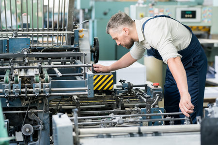 Serious concentrated bearded repairman in overall examining industrial machine and adjusting it before workの写真素材