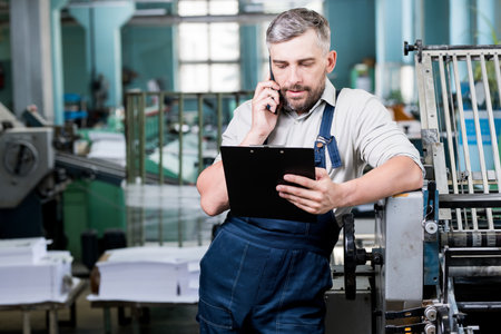 Serious confident handsome bearded foreman in workwear using cellphone while checking information in clipboardの写真素材