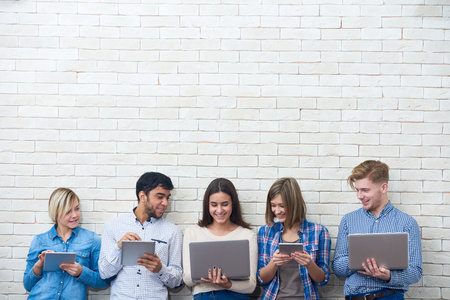 Cheerful friends watching funny video on laptop while distracted from browsing Internet with help of their gadgets, white brick wall on backgroundの写真素材