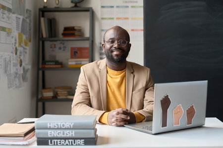 Portrait of African American teacher smiling at camera while sitting at table with laptop and books in the classroomの写真素材