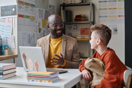 Smling African AMerican teacher using laptop to explain topic to his pupil after lessons while they sitting at tableの写真素材