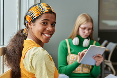Portrait of African American schoolgirl smiling at camera while talking to her classmate in the classroomの写真素材
