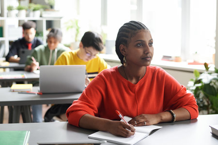 African American schoolgirl sitting at desk and making notes during lesson in the classroomの写真素材