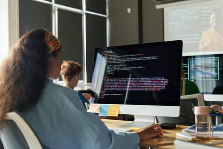 Rear view of schoolgirl sitting at desk in front of computer monitor and working with computer codesの写真素材
