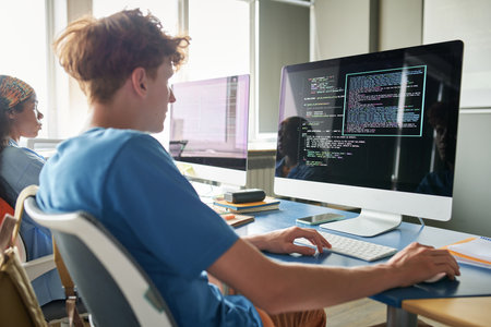 Rear view of student sitting at desk with computer and typing codes during IT lessonの写真素材