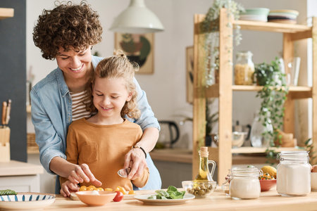 Happy young mom teaching her son to cut vegetables for salad while they cooking together in kitchenの写真素材