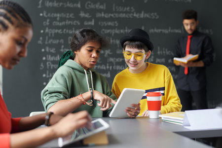 Multiethnic students discussing online material on digital tablet while sitting at table in the classroom during lessonの写真素材