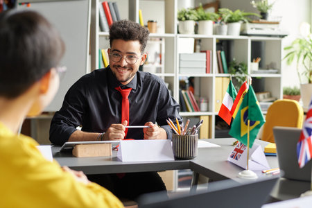 Young teacher talking to student while they sitting at table during lesson in the classroomの写真素材