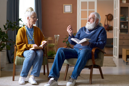 Senior couple sitting on armchairs and discussing books together during their leisure time at homeの写真素材