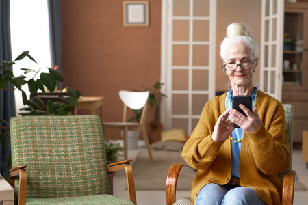 Senior woman typing message on smartphone while sitting on armchair in the living room