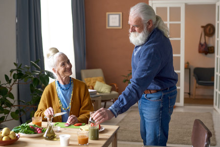 Senior man drinking fresh smoothie and talking to his wife while she cutting vegetables at tableの写真素材