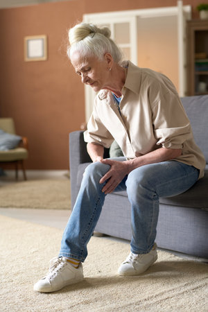 Vertical image of senior woman sitting on sofa in the living room and suffering from sore legの写真素材