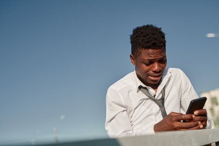 African American man standing outdoors and crying while reading shocked message on his smartphoneの写真素材
