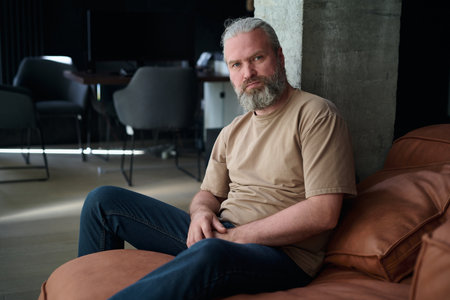 Portrait of bearded mature man looking at camera while sitting on sofa in the living roomの写真素材