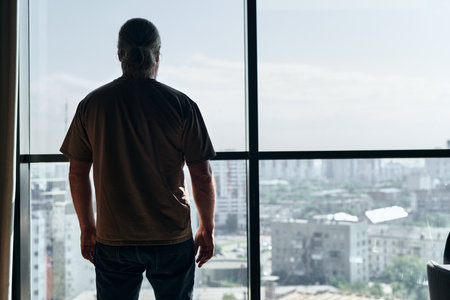 Rear view of mature man standing in front of panoramic window and looking at the cityの写真素材