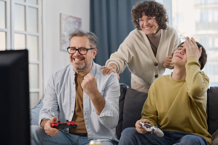Happy dad playing console game with his son while mother supporting them while they sitting on sofa at homeの写真素材