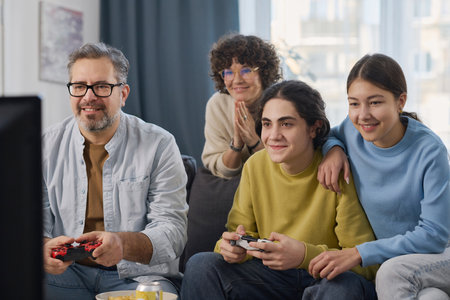 Family of four sitting on sofa in the living room and playing video game togetherの写真素材