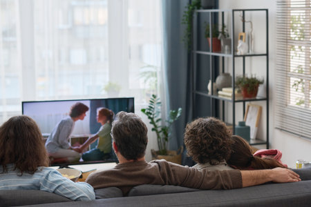 Rear view of family sitting on sofa and watching movie together at homeの写真素材