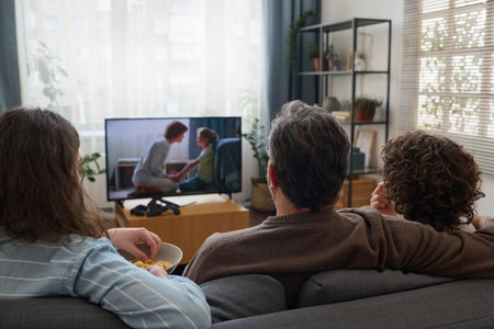 Rear view of family watching TV together in the living room while resting on sofaの写真素材