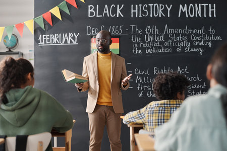 African American teacher standing against blackboard and telling about Black History Month to students in classroomの写真素材