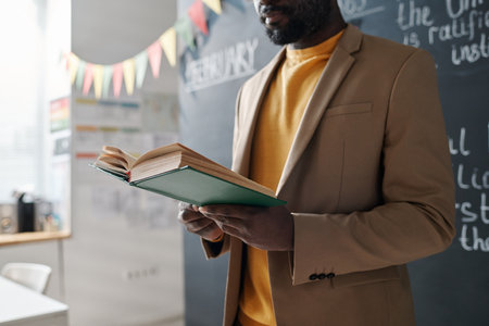 African American teacher standing near the blackboard and reading textbook during lessonの写真素材