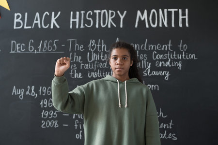 Portrait of African American schoolgirl standing against the blackboard and gesturing, she answering...