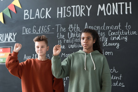Schoolboy and schoolgirl standing at blackboard and gesturing, they telling about Black History Month during a lessonの写真素材