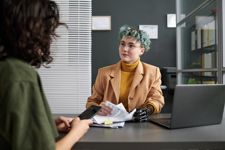Young woman with prosthesis talking to customer and discussing documents while having meeting in officeの写真素材