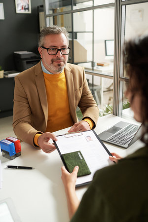 Vertical image of manager of visa center taking documents from immigrant during their meetingの写真素材