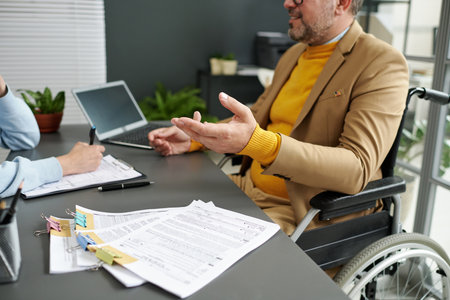 Businessman with disability sitting in wheelchair and talking to woman at meeting in officeの写真素材