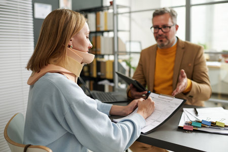 Young woman with injury of neck filing insurance documents during her meeting with manager in officeの写真素材
