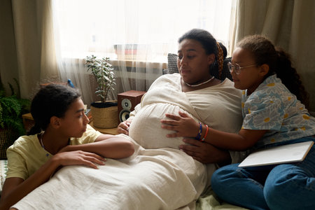 African American pregnant mom talking to her daughters during leisure time at homeの写真素材