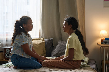 Girl talking to her elder sister while they sitting on bed opposite each other in the roomの写真素材