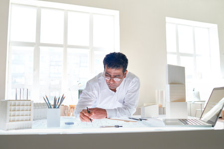 Portrait of male Indian architect drawing plans at workstation in white office interior, copy spaceの写真素材