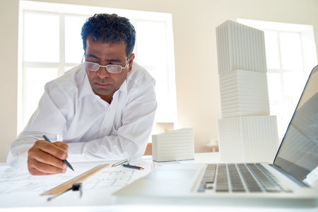 Front view portrait of mature Indian architect at workstation in office designing plans, copy spaceの写真素材
