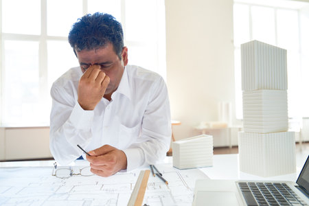Portrait of exhausted architect rubbing eyes while working at drawing table in office, copy spaceの写真素材